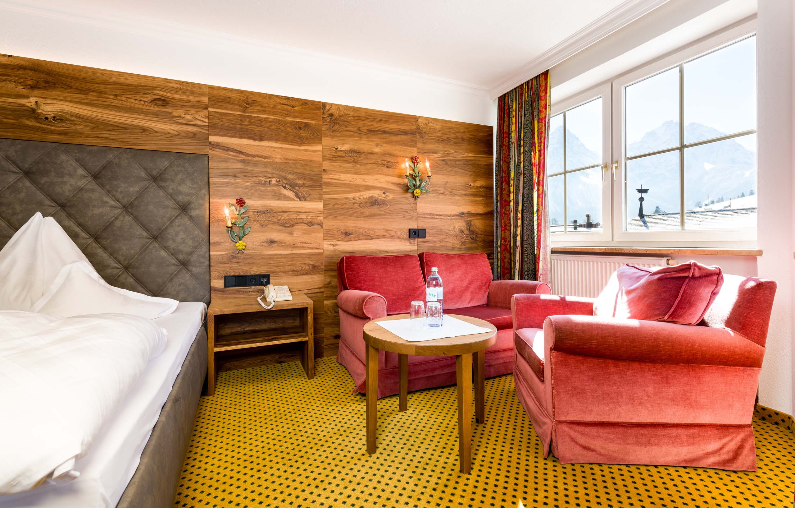 Ruime hotelkamer met gezellige zithoek, houten decoraties en uitzicht op de bergen, ideaal voor een ontspannen verblijf in de Oostenrijkse Alpen.