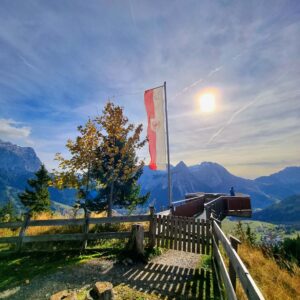 Atemberaubender Blick auf die österreichischen Alpen von einer Aussichtsplattform mit Fahne, Sommerwald, blauer Himmel und Sonne, ideal für Natur- und Wanderliebhaber.