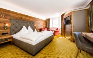 Geräumiges Hotelzimmer mit komfortablem Doppelbett, Holzdekoration, gemütlichem Sitzbereich mit Sessel und TV, perfekt für einen entspannenden Aufenthalt in den Bergen.